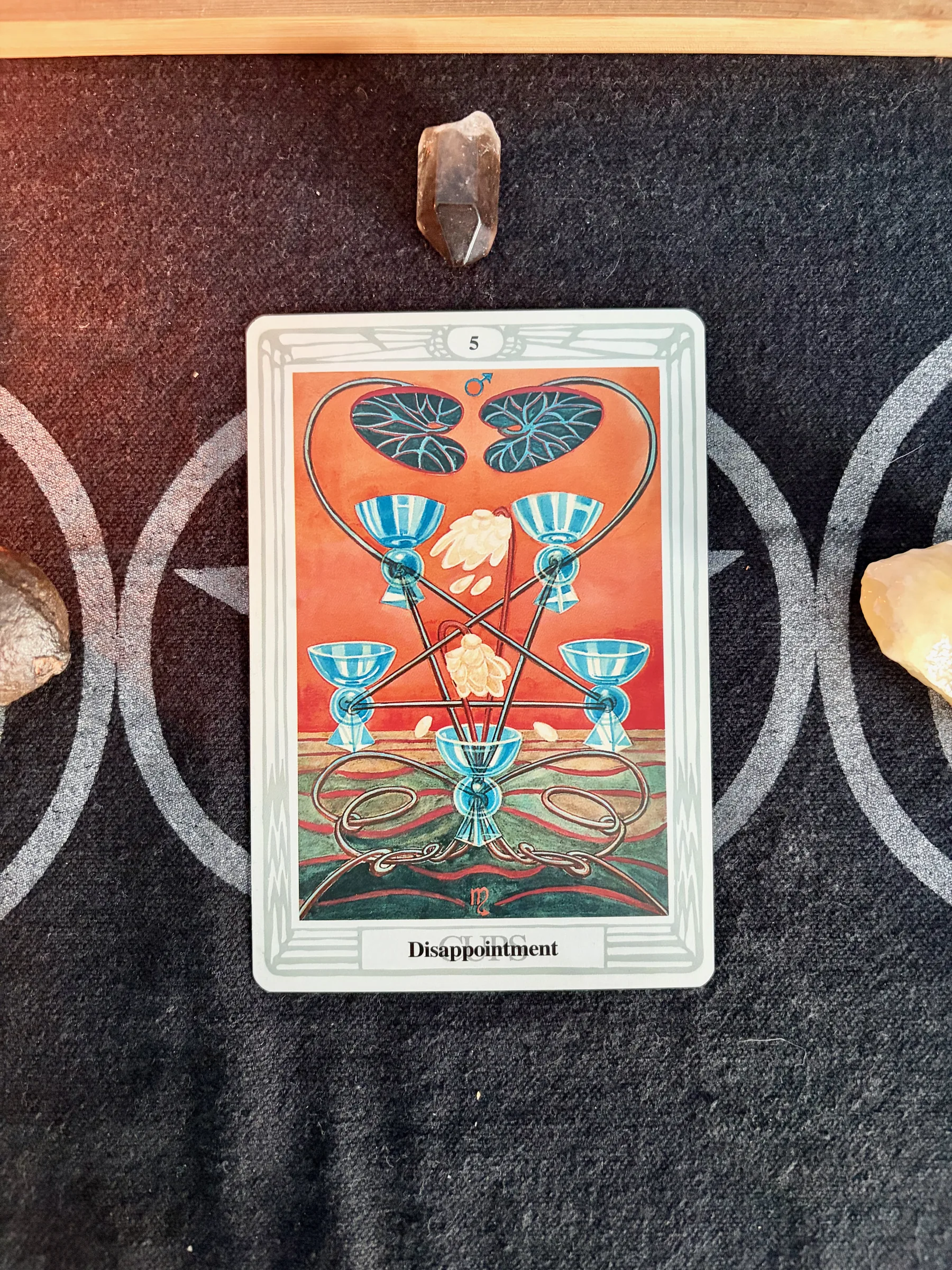 The Thoth Tarot Five of Cups, titled Disappointment, photographed on a dark patterned cloth with crystals around it.