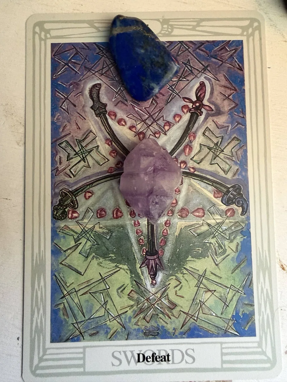 Thoth Tarot Five of Swords card, titled Defeat, with a blue stone near the top and a purple crystal over the center of the artwork.