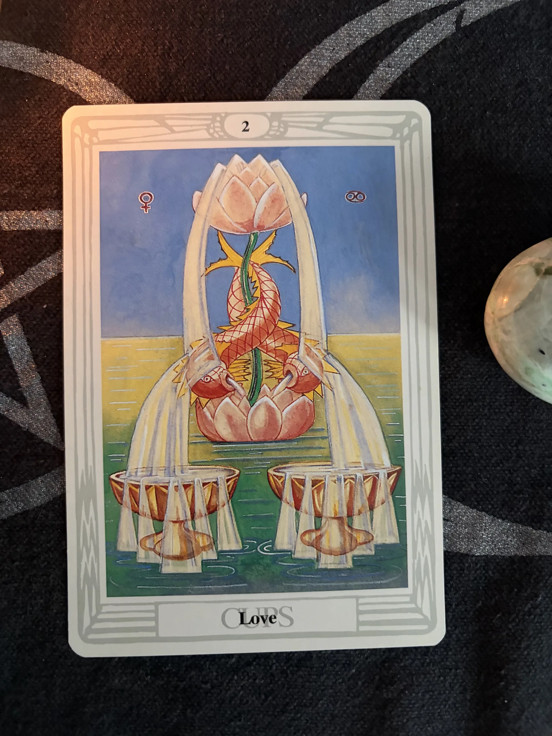 Thoth Tarot Two of Cups card, titled Love, showing two golden cups, flowing water and pink lotus flowers on a dark patterned cloth.