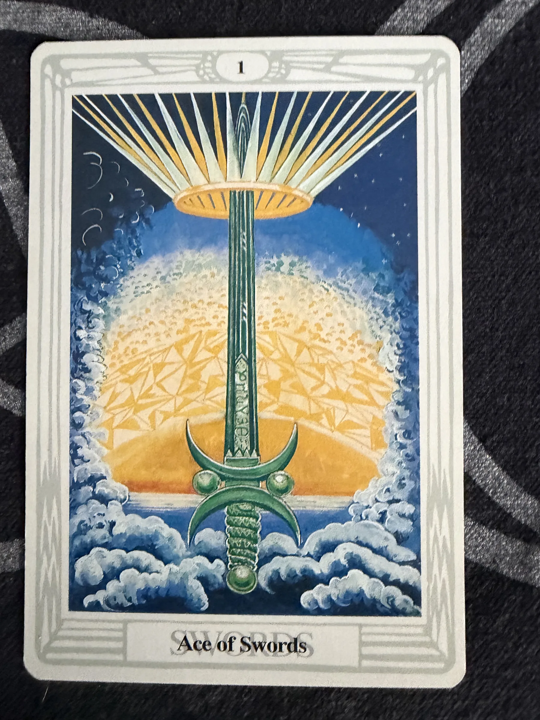 Thoth Tarot Ace of Swords photographed on a dark patterned cloth, showing an upright green sword, a golden crown, rays of light and blue-white clouds.