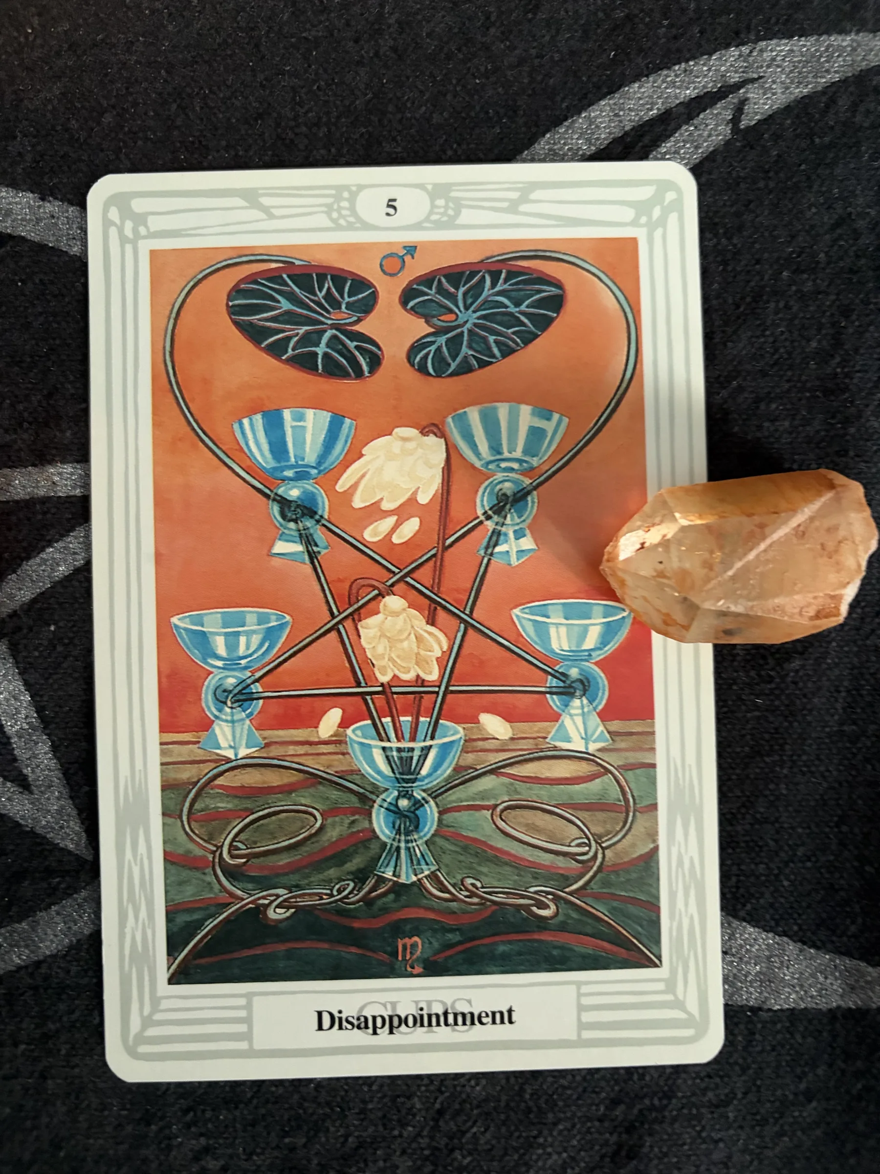 Thoth Tarot Five of Cups card, titled Disappointment, photographed on dark patterned cloth with an orange crystal resting along its right edge.