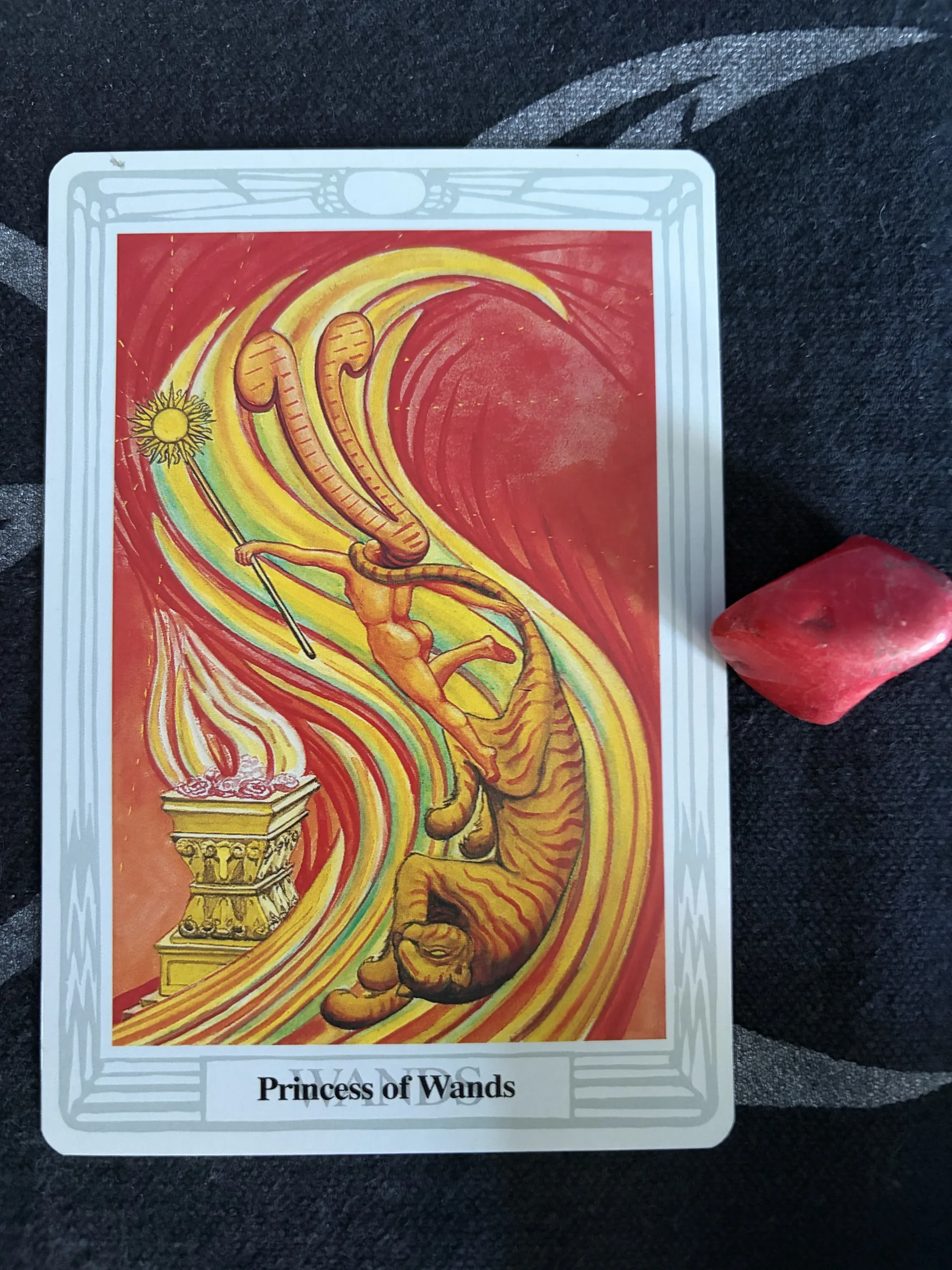 Thoth Tarot Princess of Wands card with a dancing figure, a tiger and sweeping flames, photographed on dark patterned cloth beside a polished red stone.