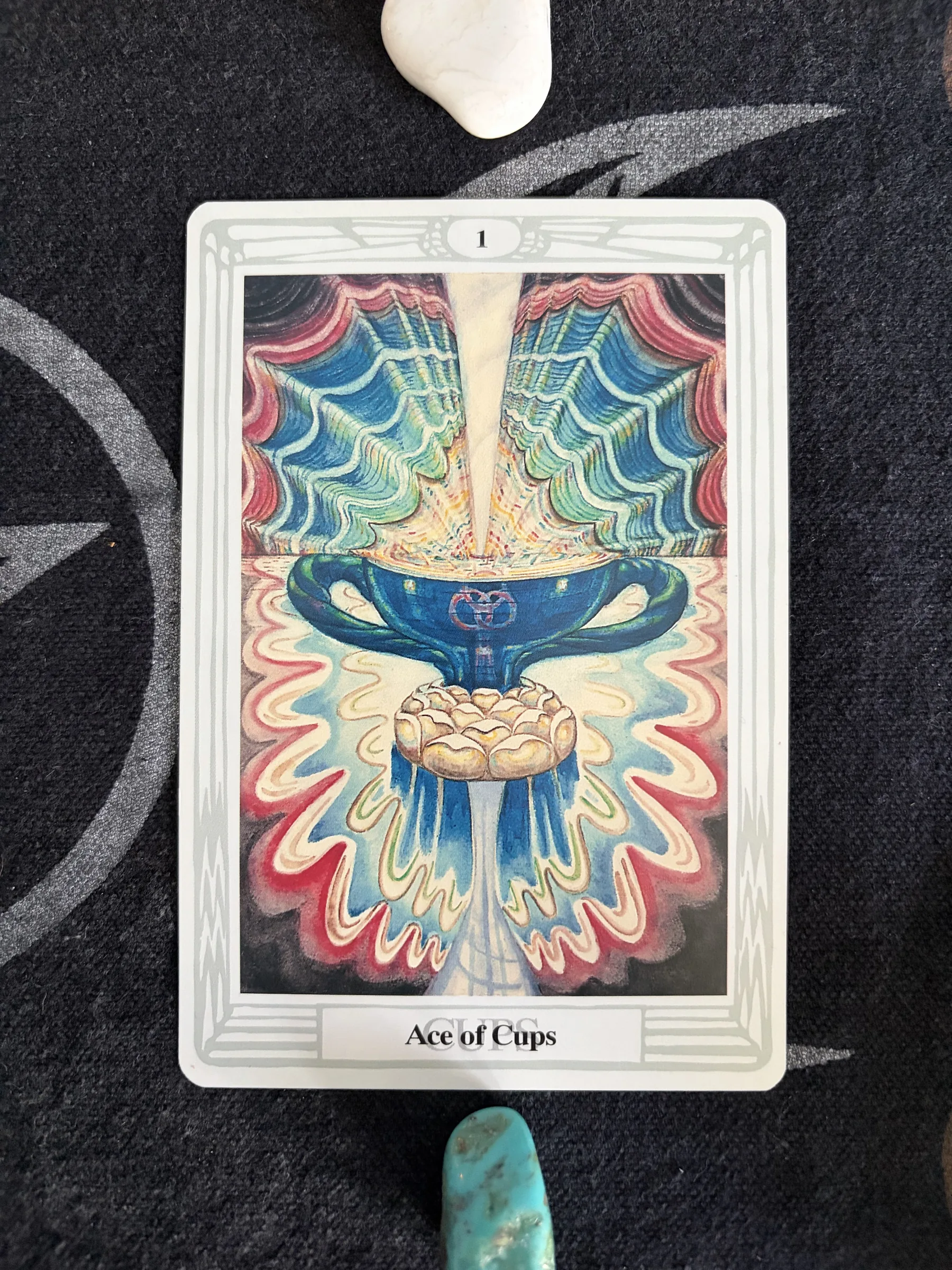 Thoth Tarot Ace of Cups card showing a blue chalice and radiant flowing patterns, photographed on dark cloth between a white stone and a turquoise-colored stone.