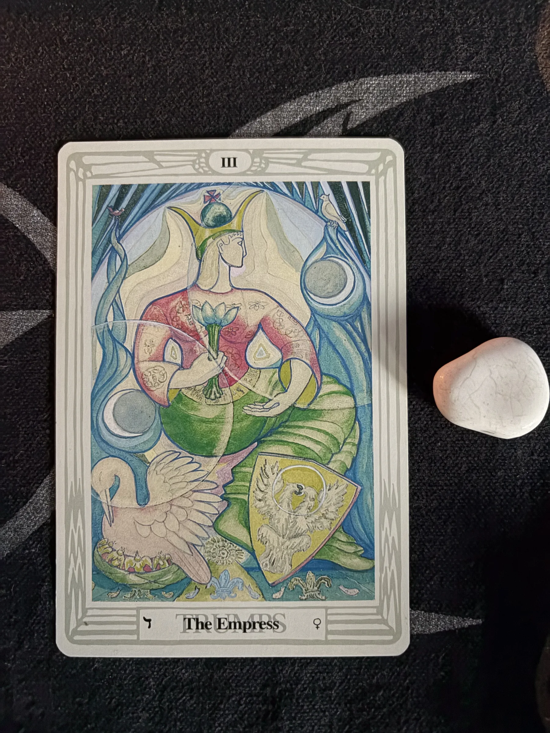 Thoth Tarot The Empress card showing a crowned figure in pink and green holding a lotus, photographed on dark patterned cloth beside a smooth white stone.