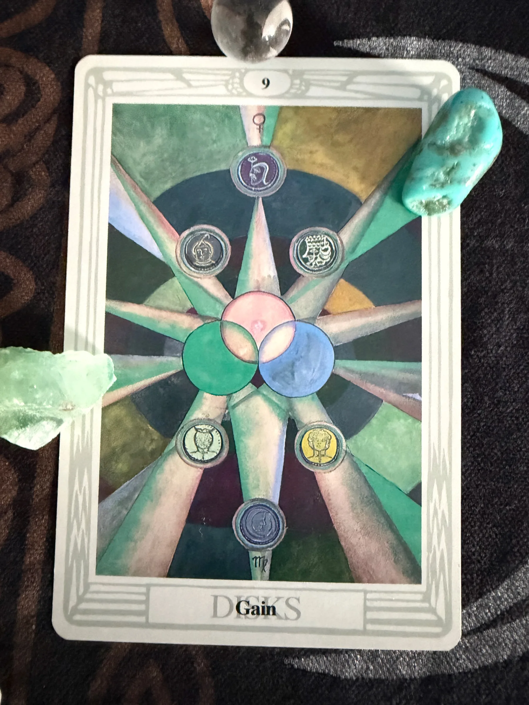 Thoth Nine of Disks Gain tarot card arranged with clear, turquoise, and pale green stones on dark patterned fabric.