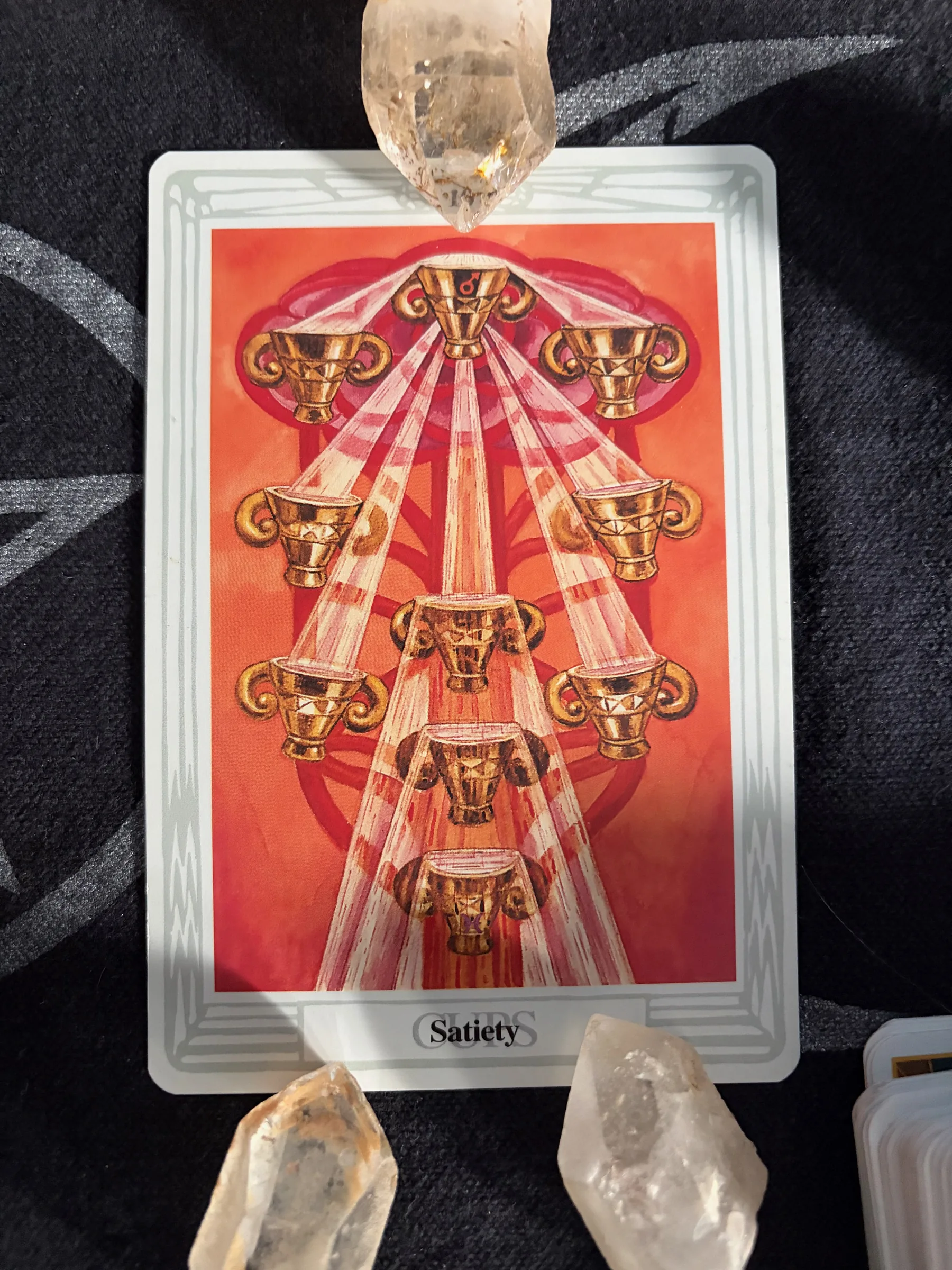 Thoth Ten of Cups Satiety tarot card arranged upright with clear quartz crystals on dark patterned fabric.