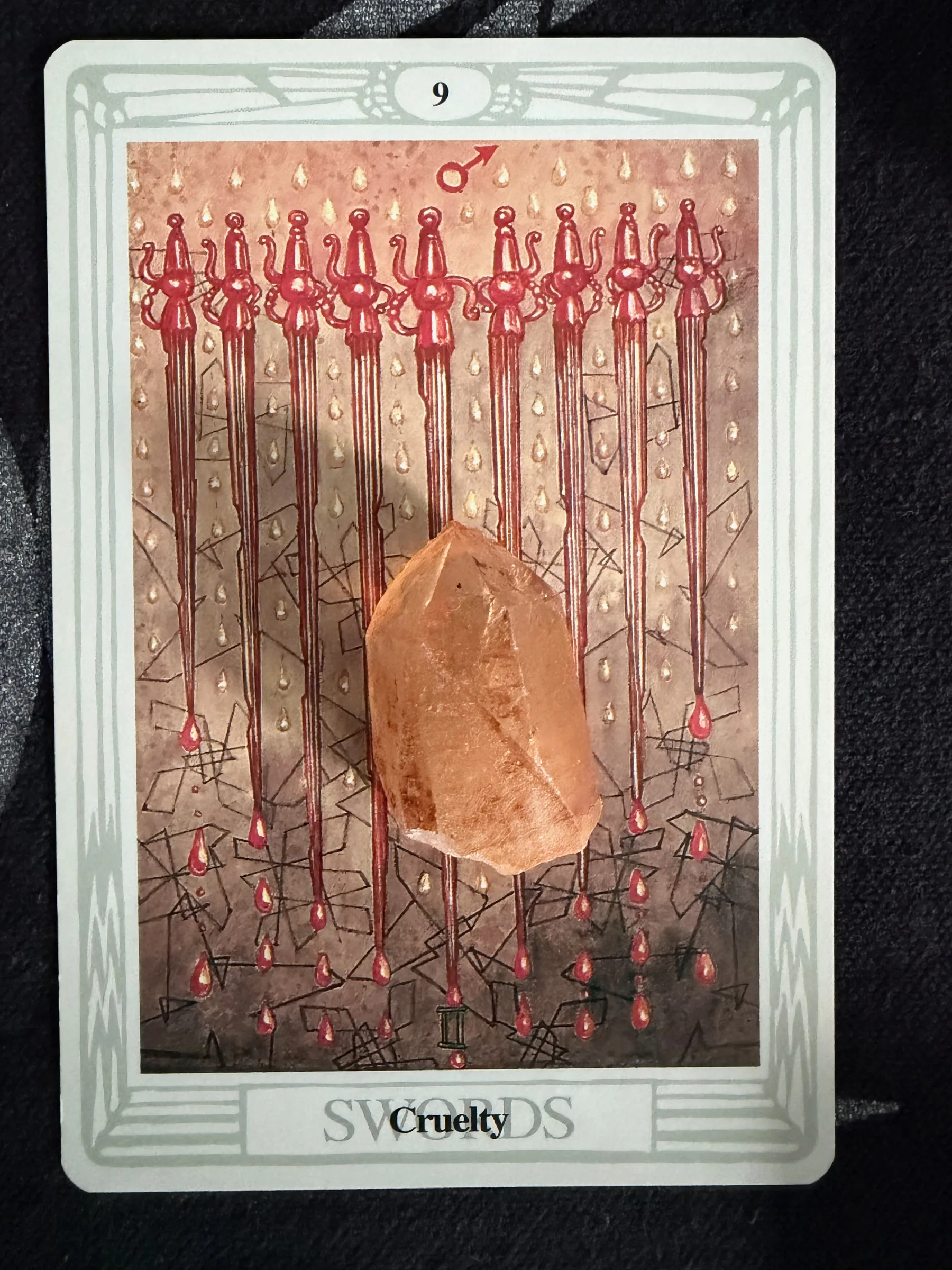 Close-up of the Thoth Nine of Swords Cruelty tarot card with nine red swords and a clear quartz crystal on dark fabric.