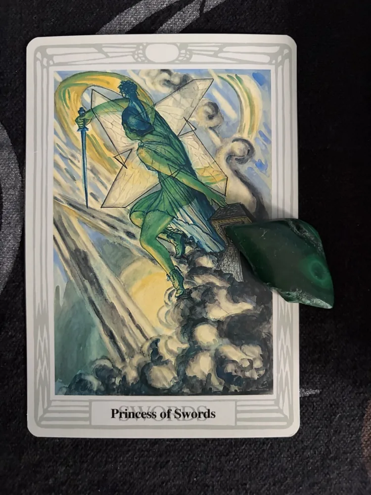 Photograph of the Thoth Princess of Swords tarot card with a polished green stone resting on its lower-right edge against dark fabric.