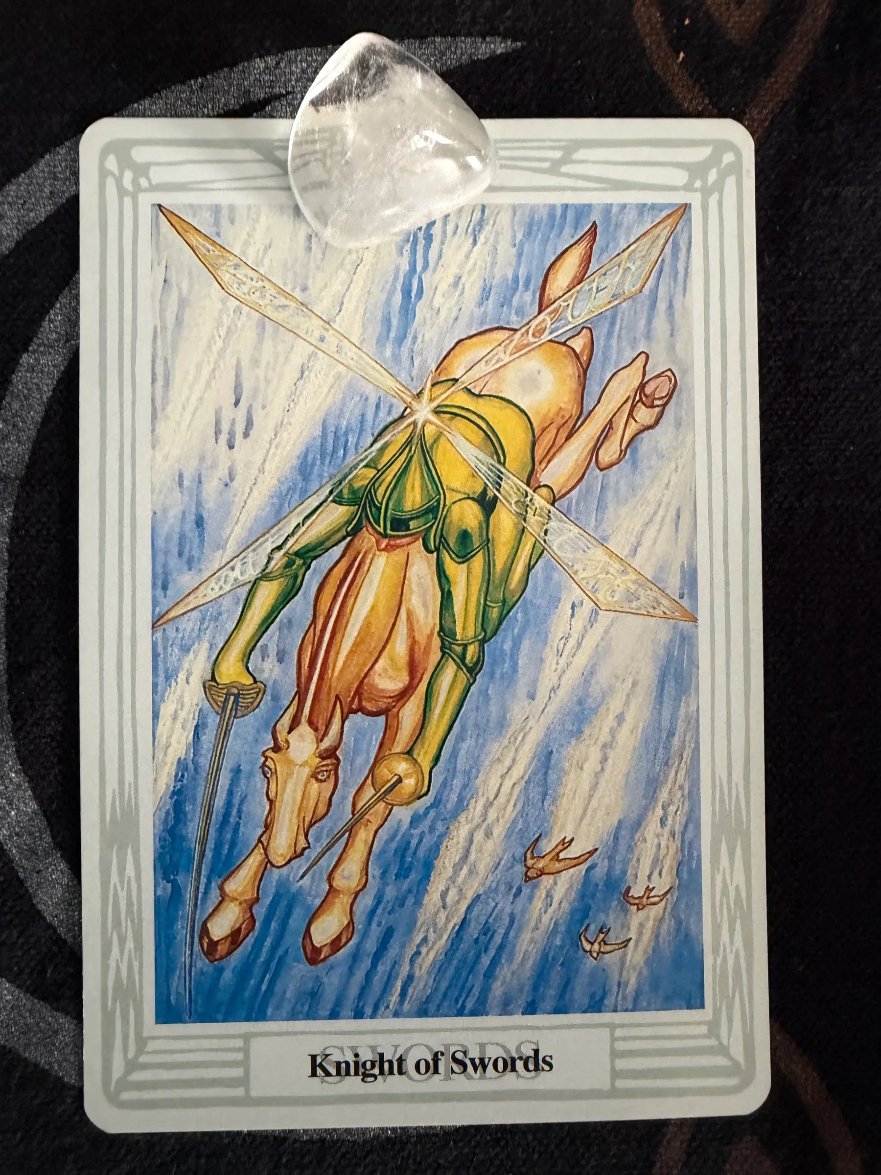 Photograph of the Thoth Knight of Swords tarot card with a clear crystal resting near the top edge against dark fabric.